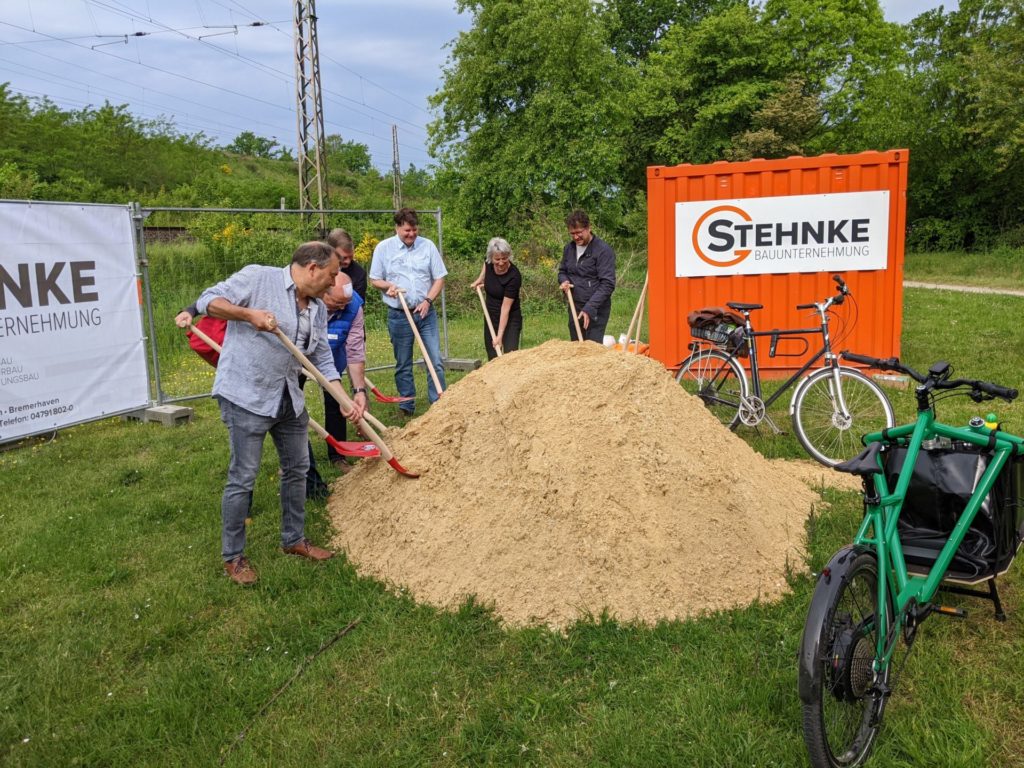 Menschen mit Schaufeln für den ersten Spatenstich vor einem Sandberg.